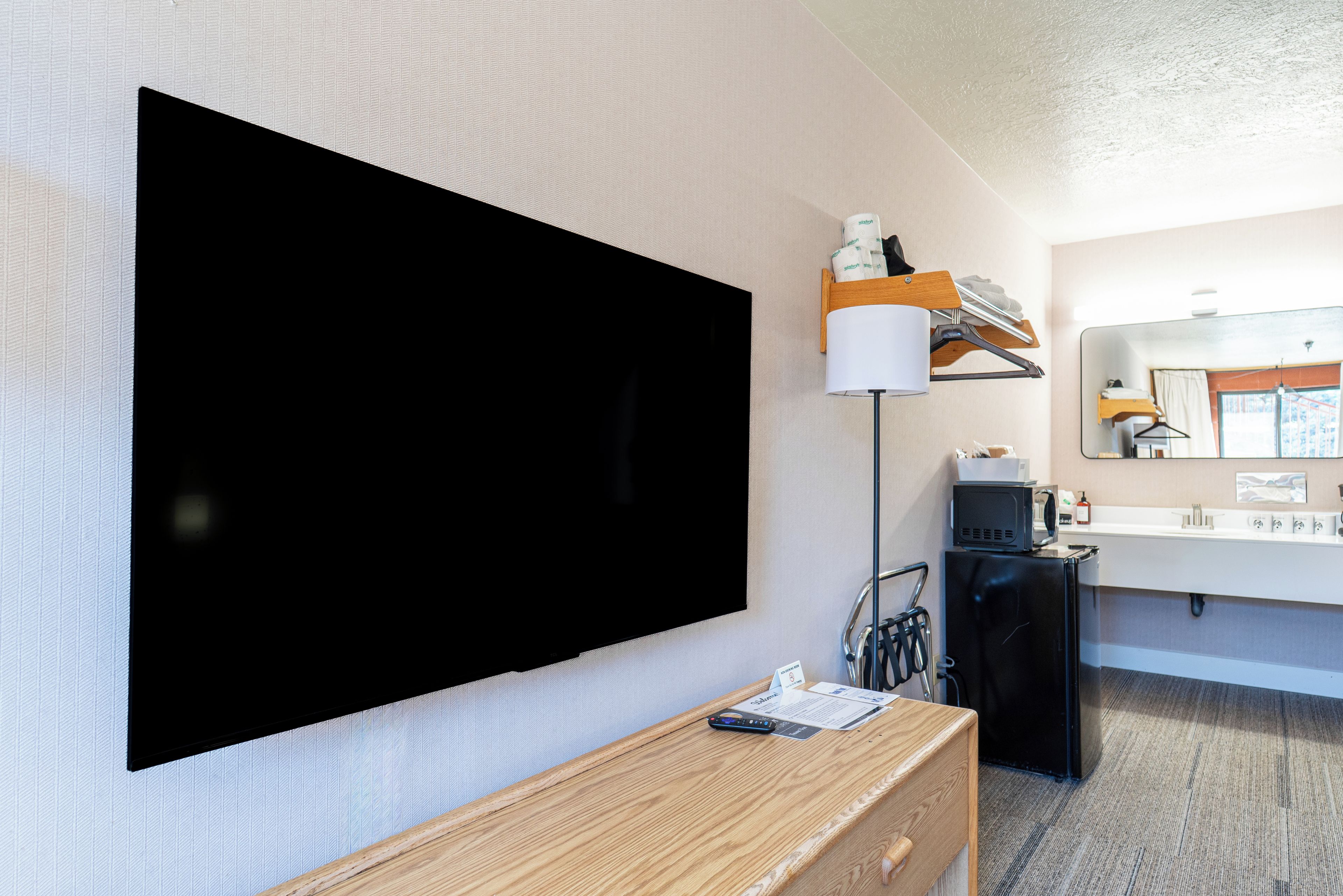Hotellihuone, jossa on suuri, seinälle asennettu taulutelevisio, puinen työpöytä, jossa on kaukosäädin, pieni musta jääkaappi, valkoinen pesuallas ja peili, jonka yläpuolella on toinen peili.