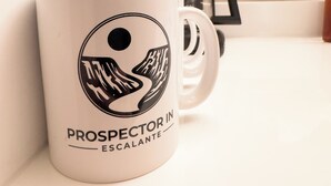 Una taza blanca con un logotipo y texto negros, una cafetera y un cuadro en un mostrador.