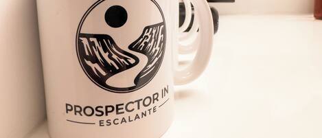 Una taza blanca con un logotipo y texto negros, una cafetera y un cuadro en un mostrador.