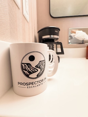 A white mug with a black logo and text, a coffee maker, and a framed picture on a counter. - Prospector Inn (Escalante)