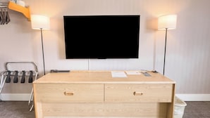 Una habitación con un televisor de pantalla plana grande montado en la pared, flanqueado por dos lámparas de pie. Hay un escritorio de madera con cajones y un pequeño cubo de basura a la derecha.