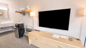 Una habitación de hotel con una televisión de pantalla plana grande montada en la pared, un aparador de madera con cajones, un cubo de basura pequeño, un espejo y un perchero para toallas.