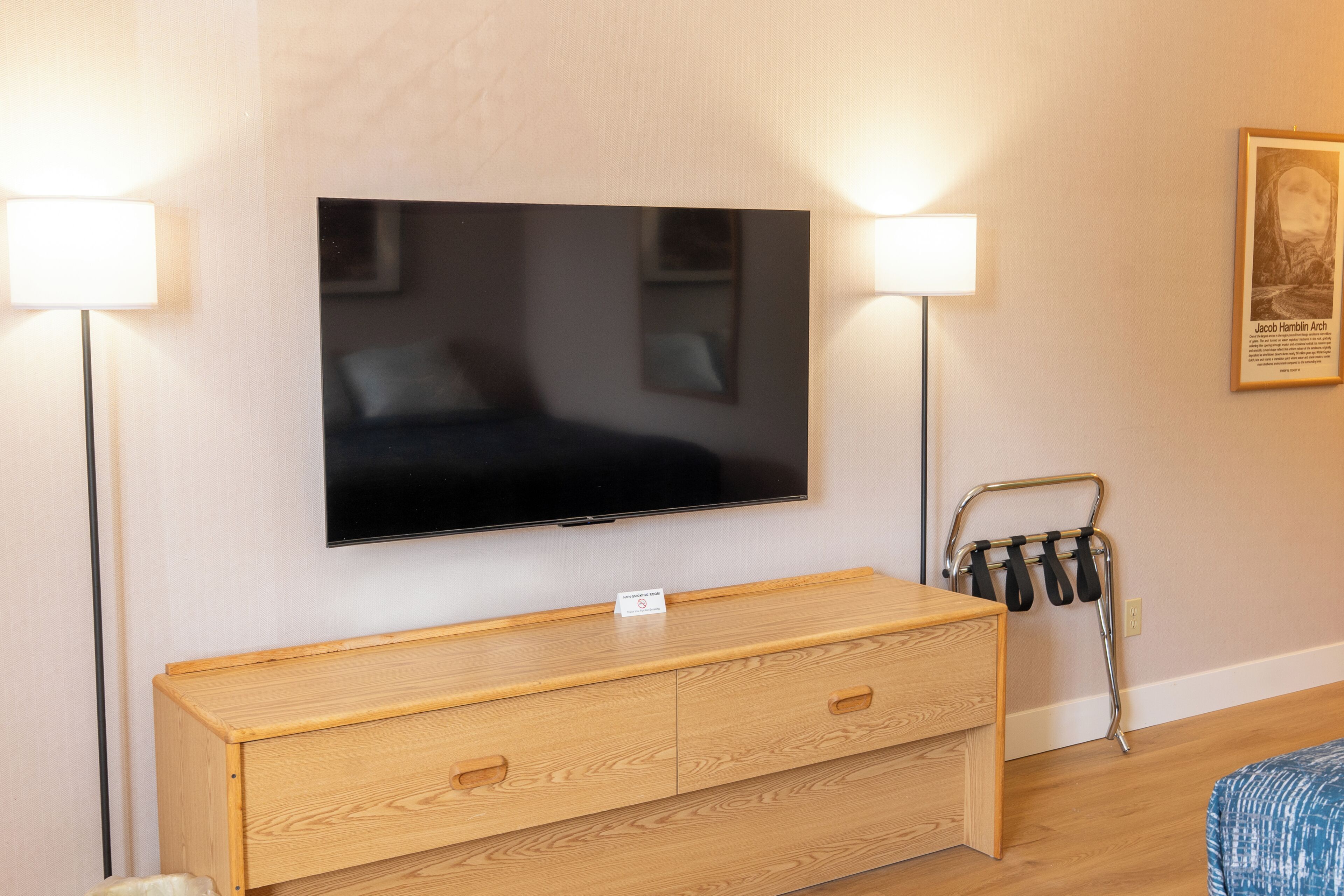 A hotel room with a flat-screen TV mounted on the wall, a wooden dresser with drawers, a metal towel rack, and a framed picture on the wall.