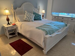 Una cama bien hecha con un cabecero blanco, dos mesitas de noche con lámparas, una alfombra roja y una ventana con persianas.
