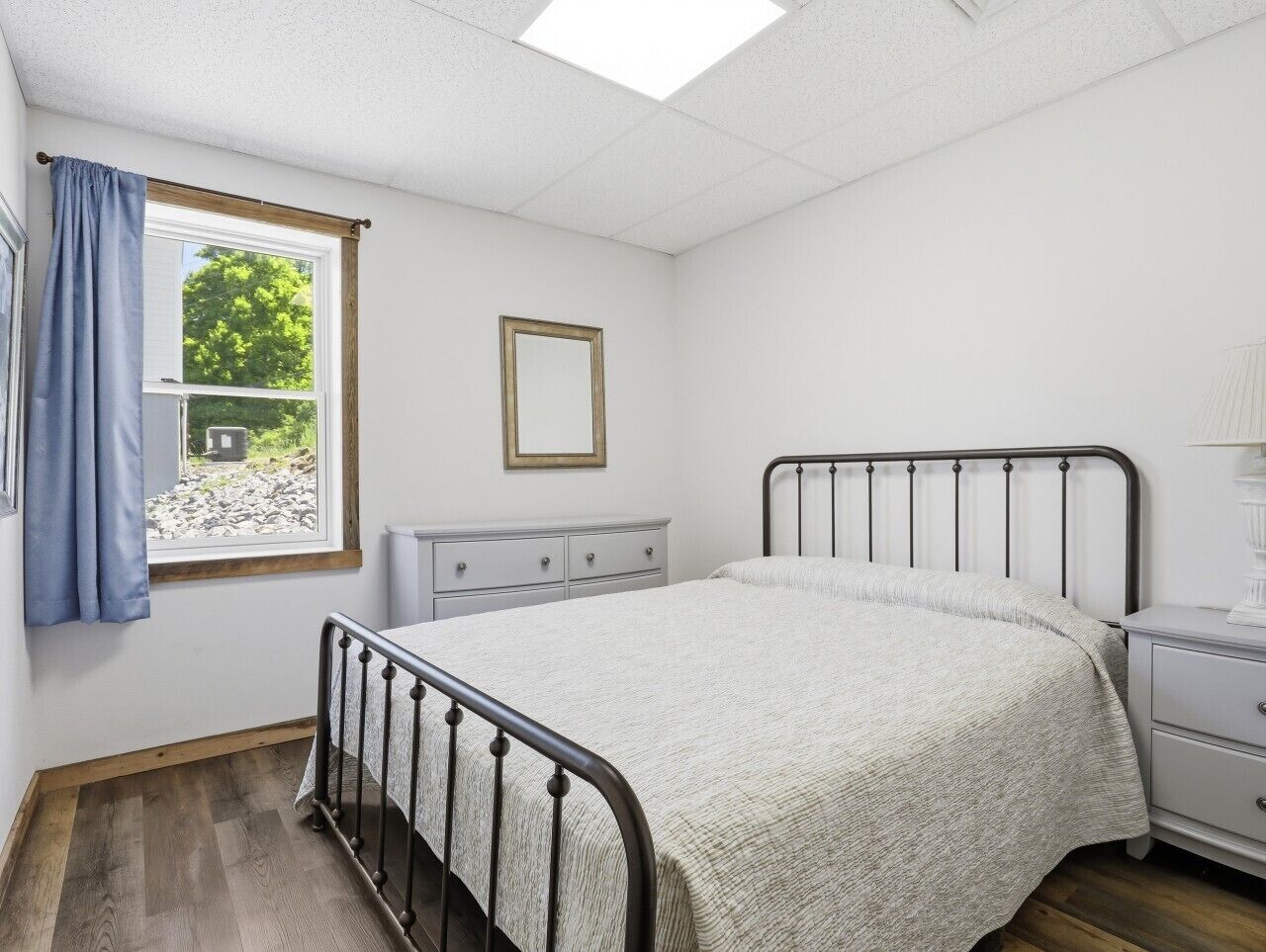 Ein Schlafzimmer mit Bett, Schminktisch, Nachttisch, einem Fenster mit Vorhängen und einem gerahmten Bild an der Wand.