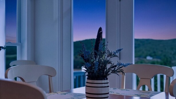 A dining table with a striped vase of flowers, white chairs, and a view of a landscape through large windows. - Lakeview Retreat Near Finger Lakes! (Lawrenceville)