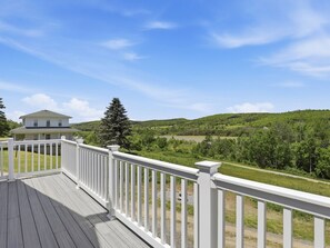 A wooden deck with white railings, a house in the background, and a view of green hills and a lake. - Lakeview Retreat Near Finger Lakes! (Lawrenceville)