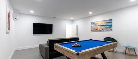 A modern room with a pool table, a flat-screen TV, a sofa, and a painting of a beach scene.