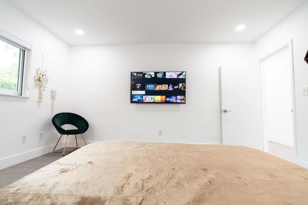 A modern room with a large wooden table, a mounted TV displaying various channels, a green chair, and a window with a view of greenery.