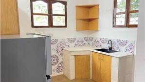 A compact kitchen with a refrigerator, a sink, and a corner cabinet. There are two windows with wooden frames and a patterned backsplash. - Spacious 4 bedroom Home with Modern Comfort Nungwi (ZANZIBAR)