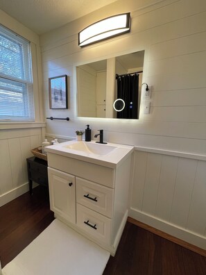A bathroom with a white vanity, a mirror, a light fixture above the mirror, and a window with blinds. - 2-bedroom house in peaceful Paragould with fenced in backyard (Paragould)