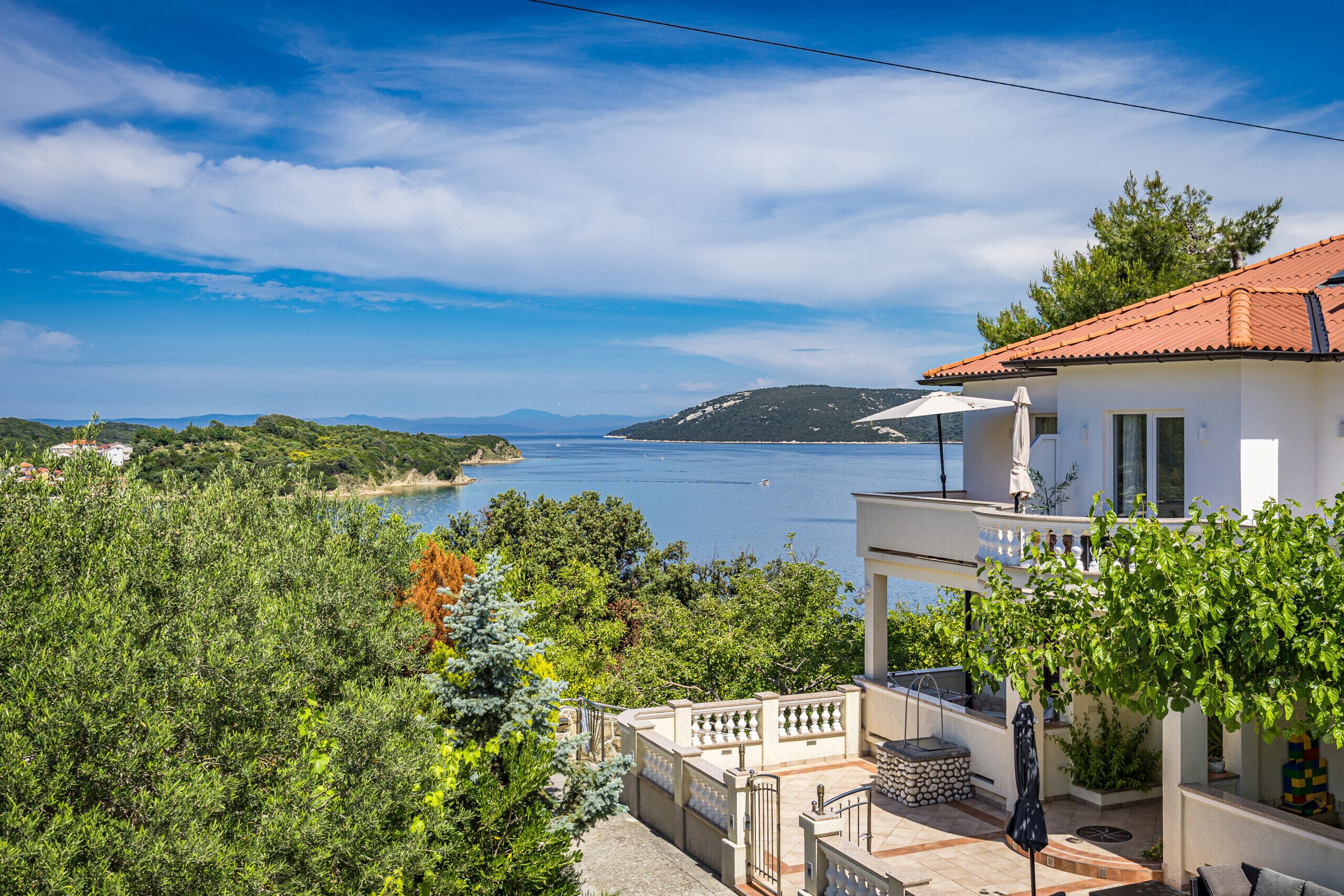 Uma propriedade costeira com varanda com vista para o mar, um edifício branco com telhado de telhas vermelhas e vegetação exuberante.