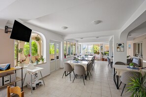 A spacious dining area with multiple tables and chairs, large windows with plants, and a television mounted on the wall.