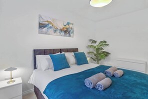Une chambre à coucher avec un lit, des oreillers, une couverture, un tableau, une plante et une lampe.