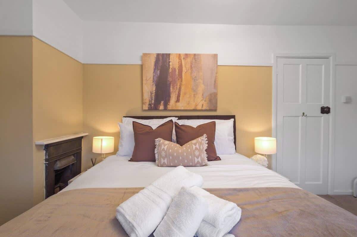 Cama bem arrumada com almofadas e um cobertor, um quadro acima da cama, uma porta à direita e uma luminária em cada lado da cama.