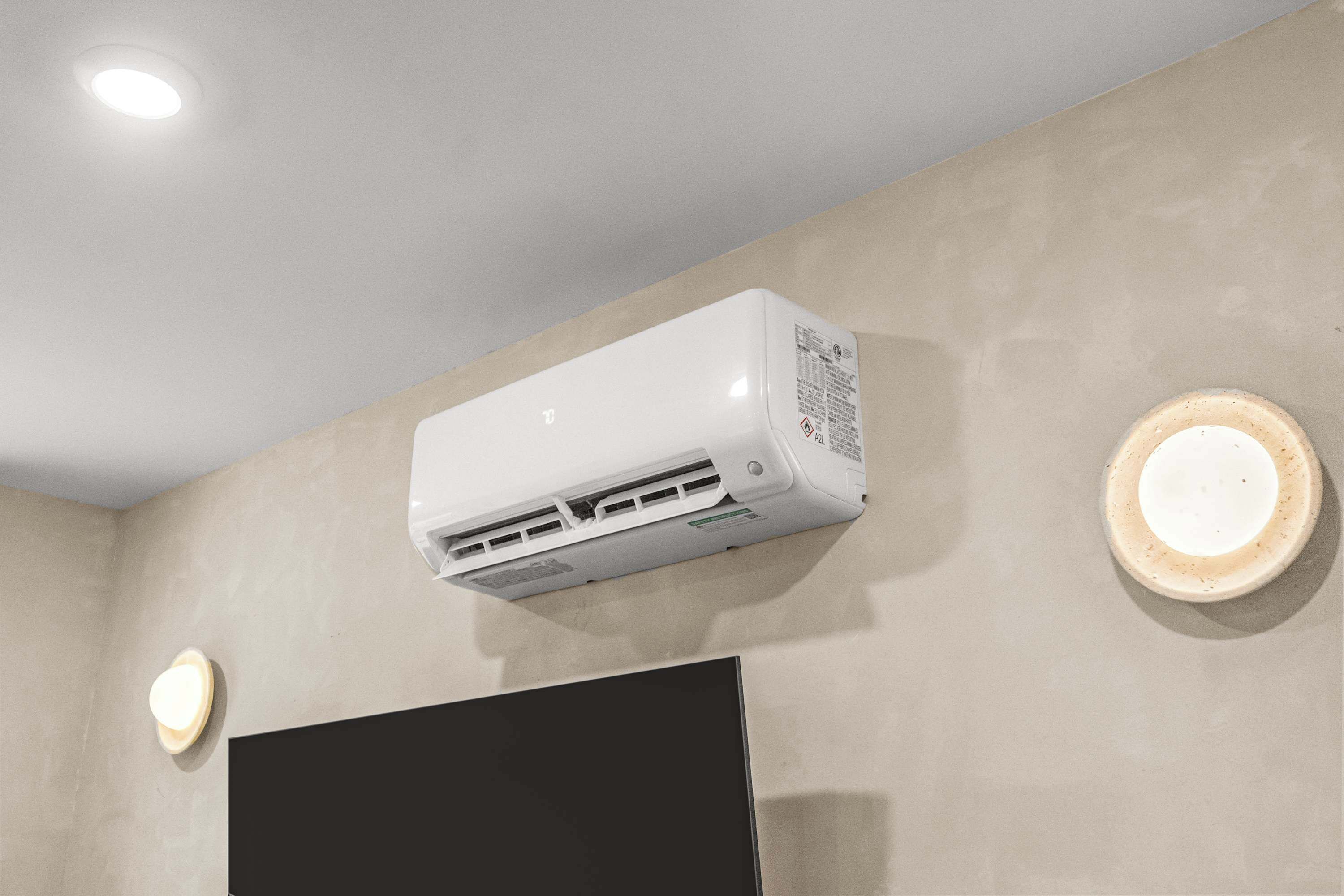 A white air conditioning unit mounted on a wall, a black flat-screen TV below it, and a round, beige decorative plate on the wall.