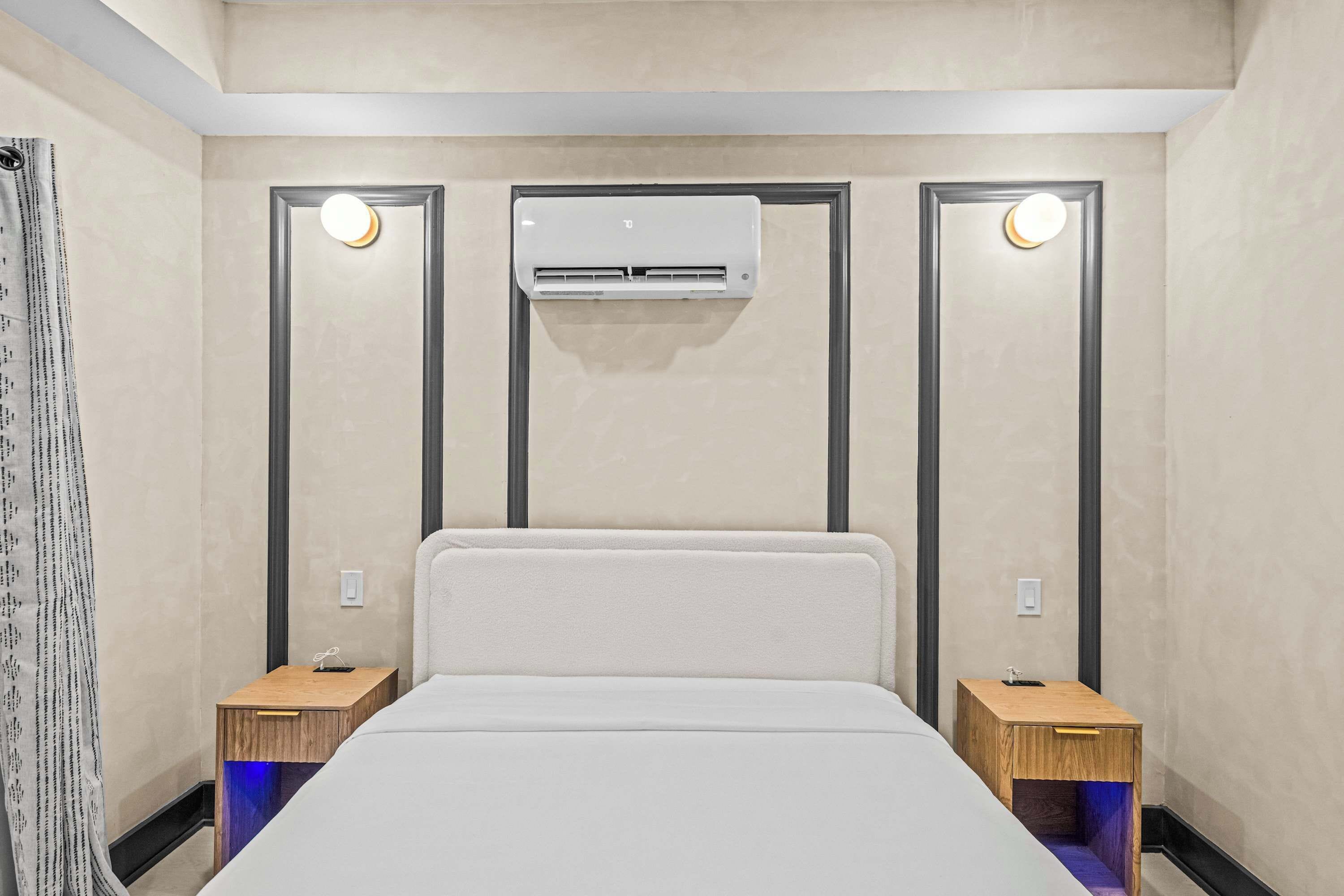 A hotel room with a bed, two bedside tables, an air conditioning unit, and a wall-mounted light fixture.