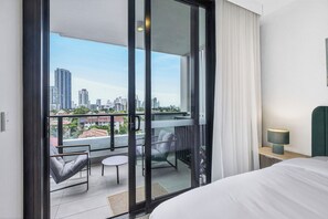 A hotel room with a balcony, a bed, a chair, a small table, and a nightstand with a lamp. - Southport 2BR Retreat | Rooftop Infinity Pool (Southport)