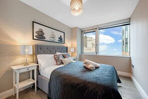 A neatly made bed with a dark quilt, a bedside lamp, and a framed artwork on the wall. - Brighton's Beachfront Gem! | The Lanes | Sleeps 6 (Brighton and Hove)