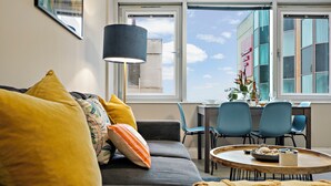 Een moderne woonkamer met een grijze bank, gele en meerkleurige kussens, een ronde houten salontafel, en een eethoek met blauwe stoelen en een tafel.