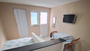 A small, compact room with a bed, a desk, a chair, a TV mounted on the wall, and a window with blinds.