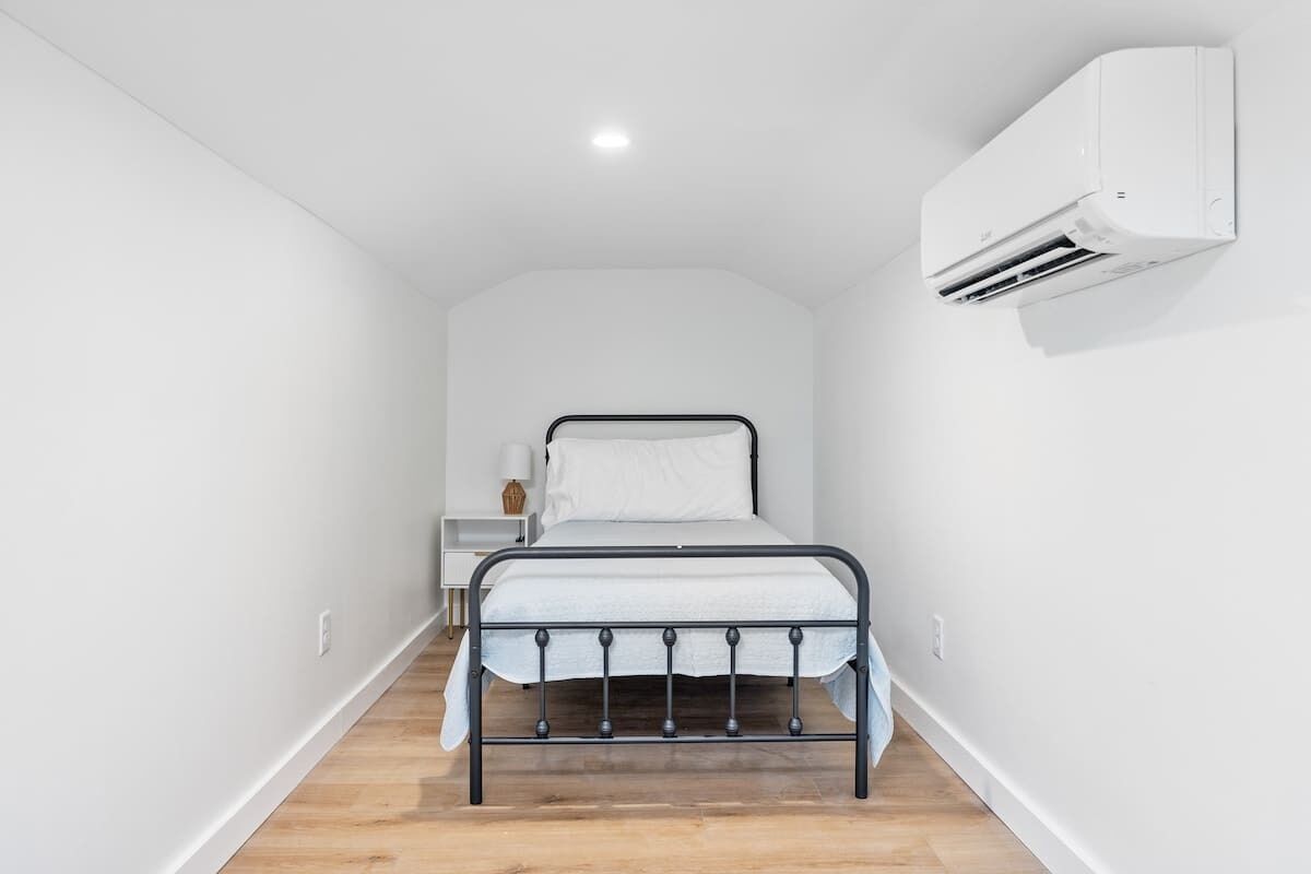 Quarto estreito, com paredes brancas, uma cama, uma mesinha de cabeceira e ar condicionado fixado na parede.