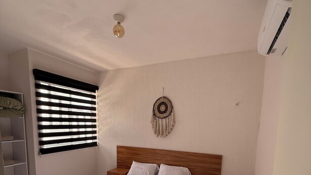 A bedroom with a wooden bed, white bedding, a wall hanging, and a window with blinds. - Tranquil 2-bedroom apartment with AC in welcoming Tulum (Tulum)