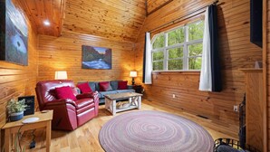Interior de una cabaña de madera con un sofá de cuero, una alfombra redonda, un cuadro y una ventana con cortinas.