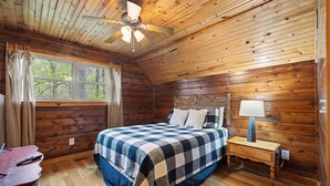 Un dormitorio de cabaña de madera con cama, mesita de noche, lámpara y ventilador de techo.