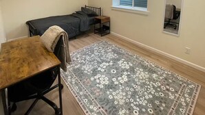 A small, well-lit room with a bed, a desk, a chair, a rug, and a window with blinds. - Charming brand new home! Walk to everything! (Atchison)