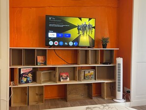 A living room with a mounted TV displaying a streaming service interface, wooden shelves with books, and a standing fan. - Grand T2 Cosy -Rouen historic center near Hôtel de Ville - L'Hauss'Rouennais (Rouen)