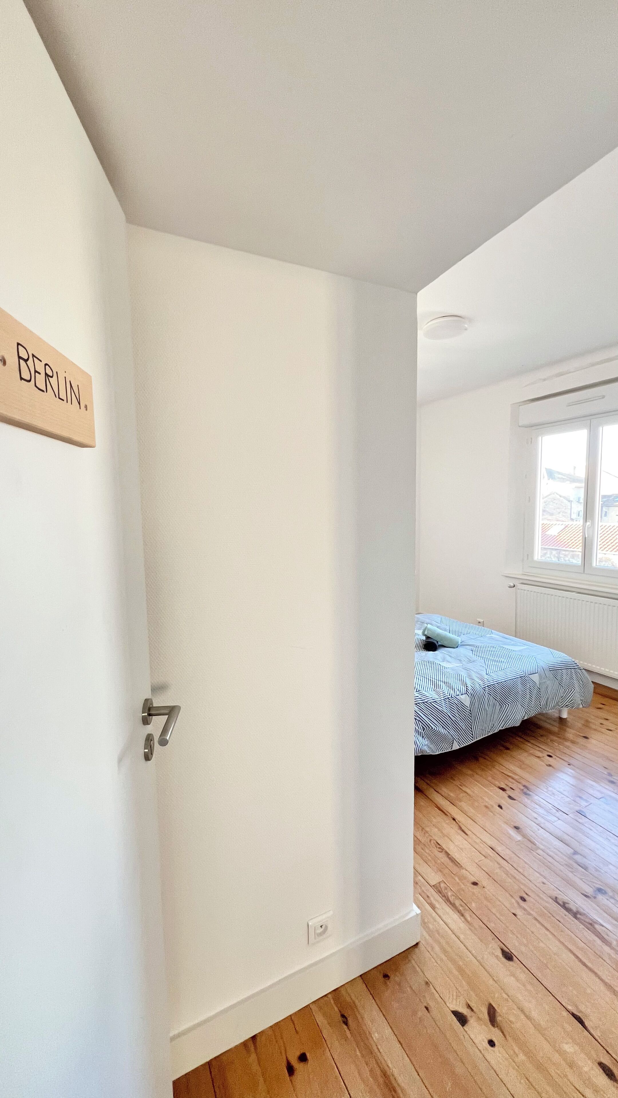 Une pièce avec un plancher en bois, un lit recouvert d’une courtepointe à rayures, une fenêtre donnant sur des immeubles et une porte sur laquelle est inscrit le mot « BERLIN ».