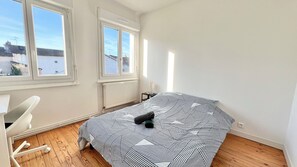 Ein Schlafzimmer mit einem Bett, einem Schreibtisch und einem Sessel. Zwei Fenster und eine Heizung sind vorhanden.