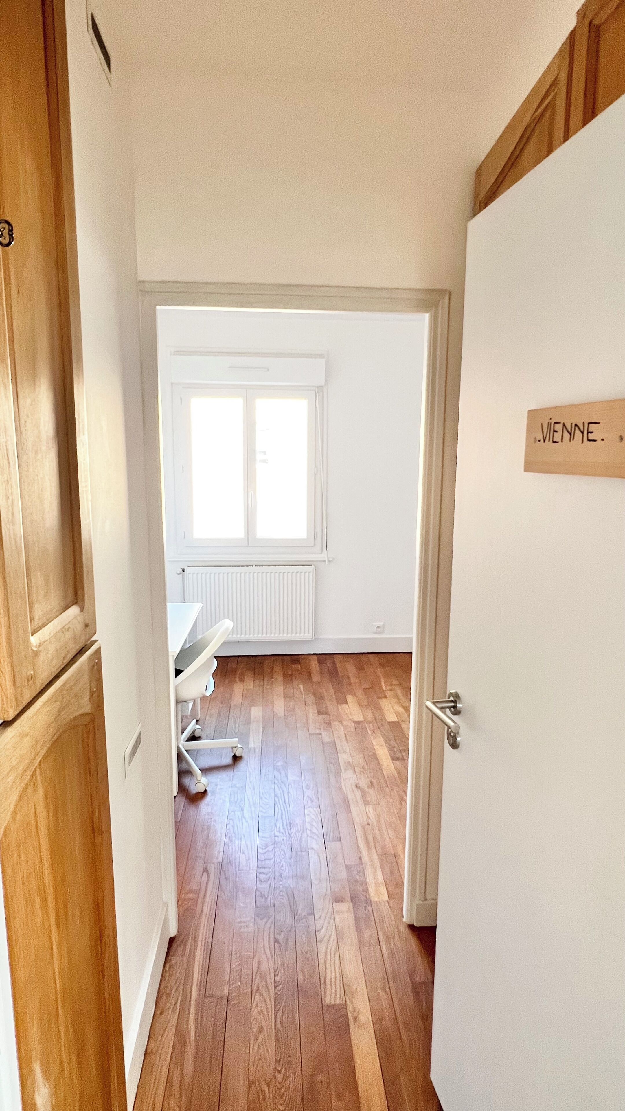 Una habitación con suelo de madera, un radiador blanco, una ventana con persianas y una puerta con la leyenda 'VIENNE'.