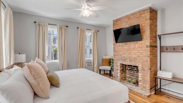 Ein Schlafzimmer mit Kamin aus Ziegelsteinen, einem Bett mit Kissen, einem Deckenventilator und einem an der Wand befestigten Fernseher.