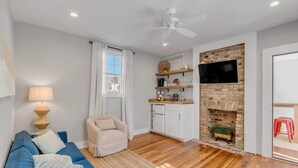 Ein modernes Wohnzimmer mit einem blauen Sofa, einem weißen Sessel, Holzboden, Deckenventilator, Küchenbereich mit Kamin und einem an der Wand montierten Fernseher.