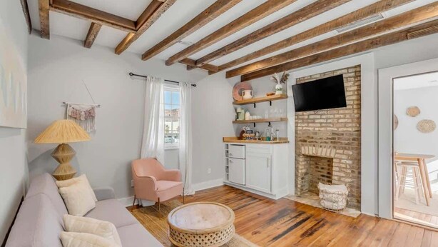 Un salon avec un canapé, une table basse ronde et un fauteuil rose. On y trouve une cheminée en briques, un téléviseur fixé au mur et une fenêtre avec des rideaux.