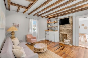A living room with a sofa, round coffee table, and a pink chair. There is a brick fireplace, a television mounted on the wall, and a window with curtains.