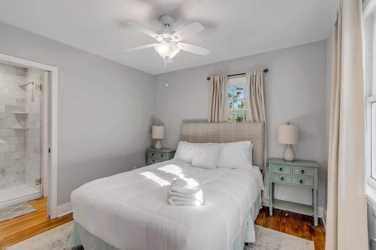 A bedroom with a bed, two bedside tables with lamps, a ceiling fan, a window with curtains, and a glass shower enclosure.