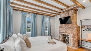 Une chambre avec une cheminée en briques, un lit avec une literie blanche et un téléviseur fixé au mur.
