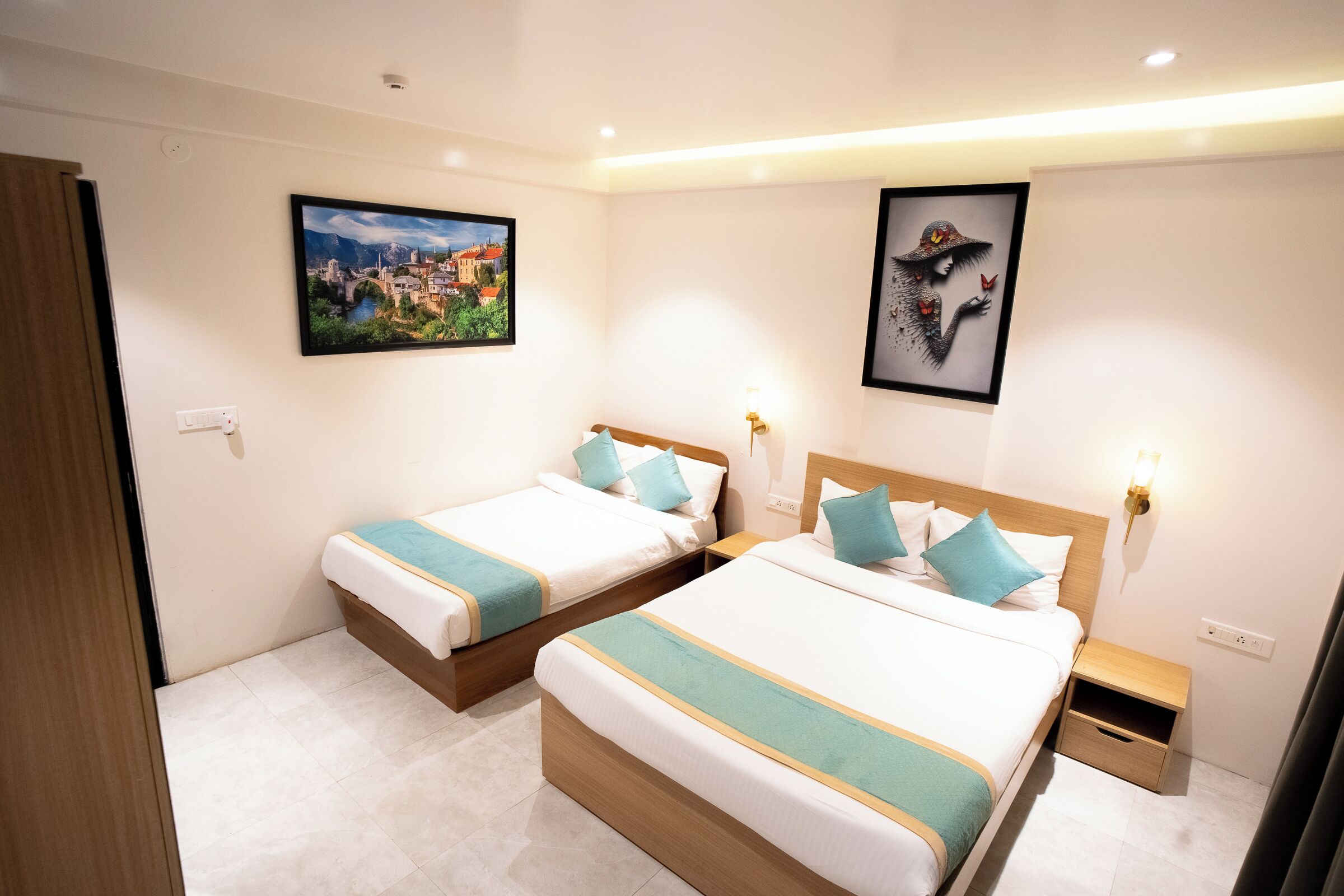 A hotel room with two beds, a nightstand, a wall-mounted light, and two framed pictures.