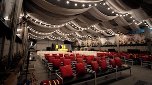 A banquet hall with rows of white chairs, red cushions, and a ceiling adorned with draped fabric and string lights. - Everest Inn Chakan (Chakan)