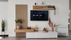 Een moderne woonkamer met een flatscreen televisie aan een houten wand, een witte tv-meubel eronder en een potplant aan de linkerkant.