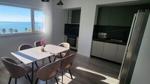 Een eetgedeelte met een tafel en stoelen, uitzicht op zee door een raam, en een keuken met moderne apparatuur.