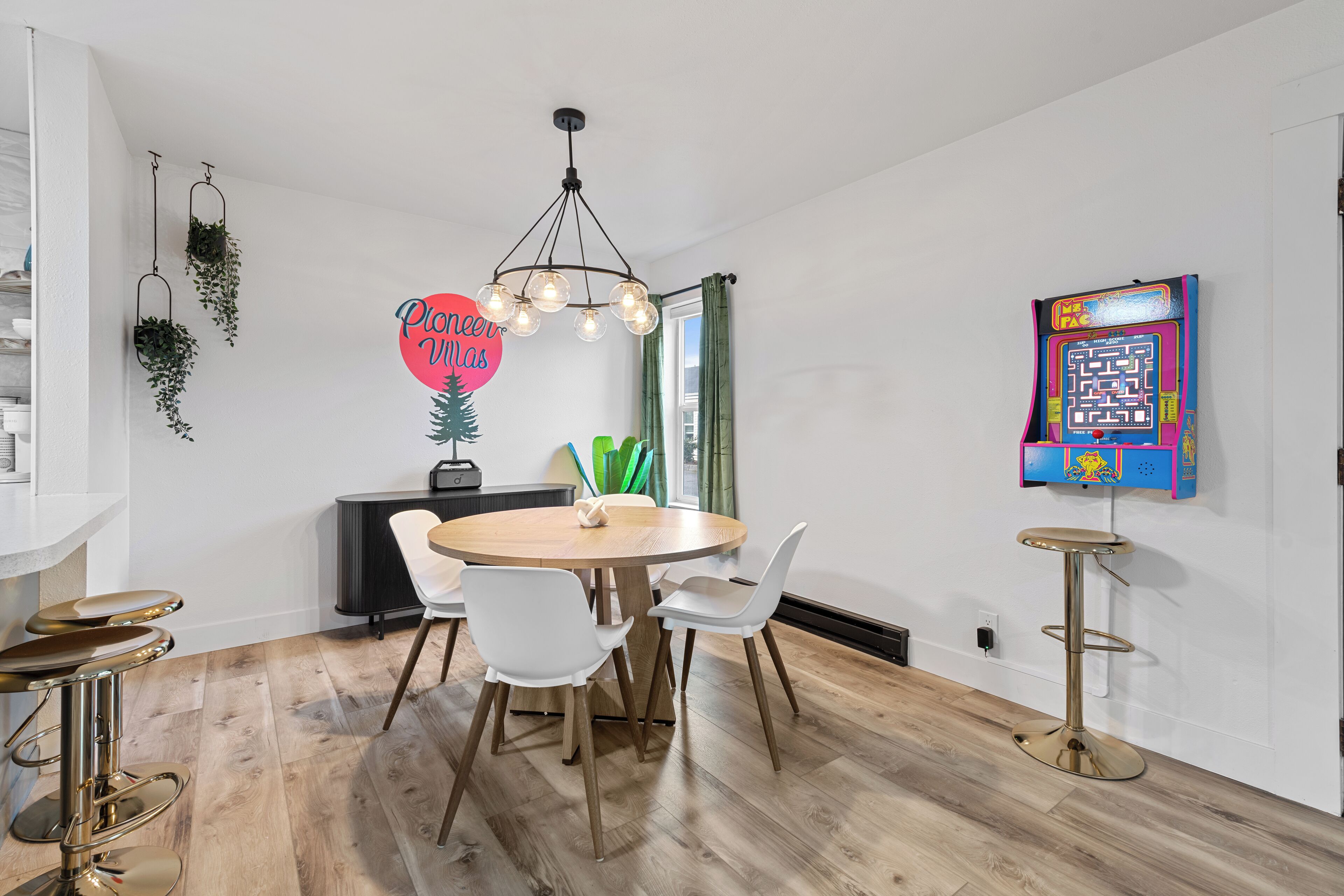 A modern dining area with a round wooden table, white chairs, hanging plants, a pendant light, and a colorful arcade game.