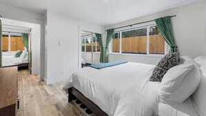 A bedroom with a bed, a wooden floor, a window with curtains, and a mirror.