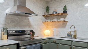 A modern kitchen with a stainless steel range, a built-in oven, and a wall-mounted extractor hood. There are wooden shelves above the sink holding dishes and a potted plant.