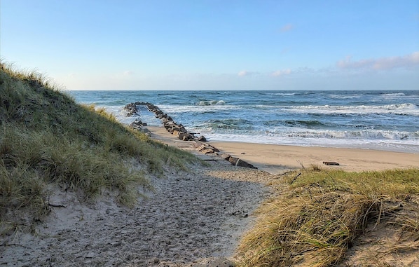 A beach path leading to the ocean, with a rocky formation on the left and waves crashing onto the shore.