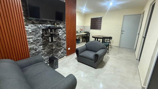 A living room with two grey sofas, a flat-screen TV mounted on a stone wall, a kitchen area with a microwave and a small table, and a door leading to another room. - Casa Villas Amanecer only 5 minutes away from Merida's periferico (Kanasín)