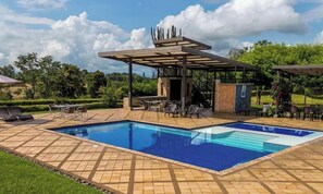 Ein rechteckiger Pool mit einer gefliesten Liegefläche, einer überdachten Terrasse mit Stühlen und einer Backstein-Struktur.